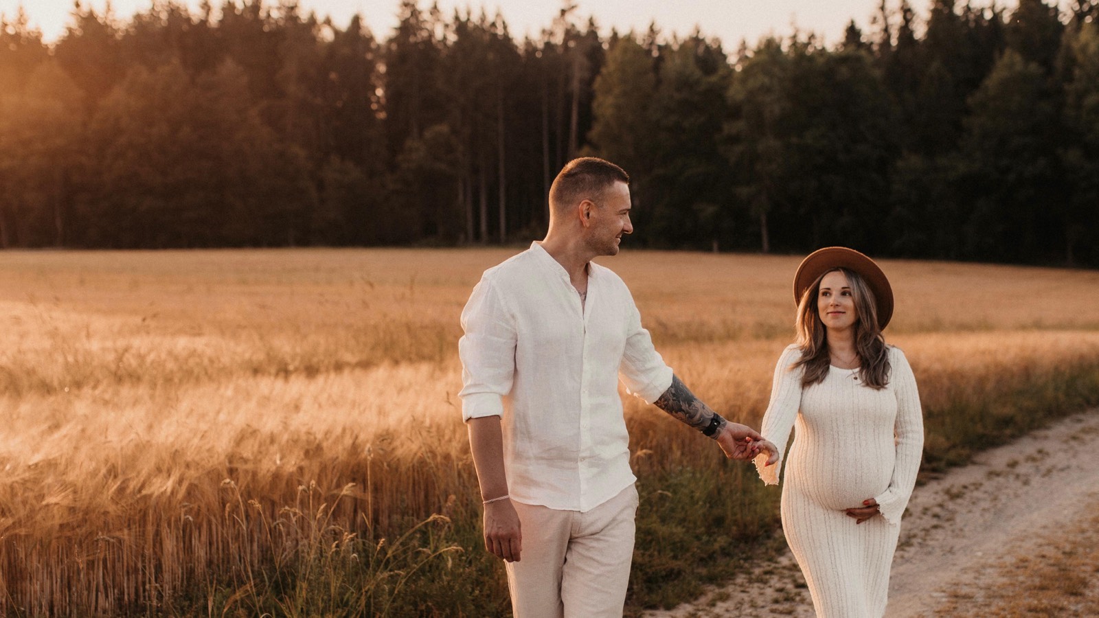 Schwangere im weißen Kleid mit Hut geht Hand in Hand mit ihrem Partner einen Feldweg am Getreidefeld entlang in der Abendsonne -- Babybauch-Shooting von Simple Moments