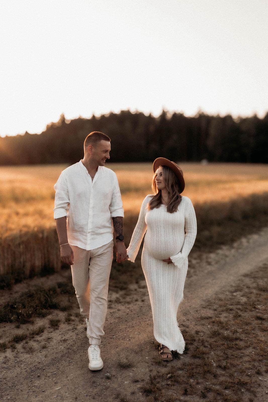 Schwangeres Paar spaziert Hand in Hand über einen Feldweg im Abendlicht