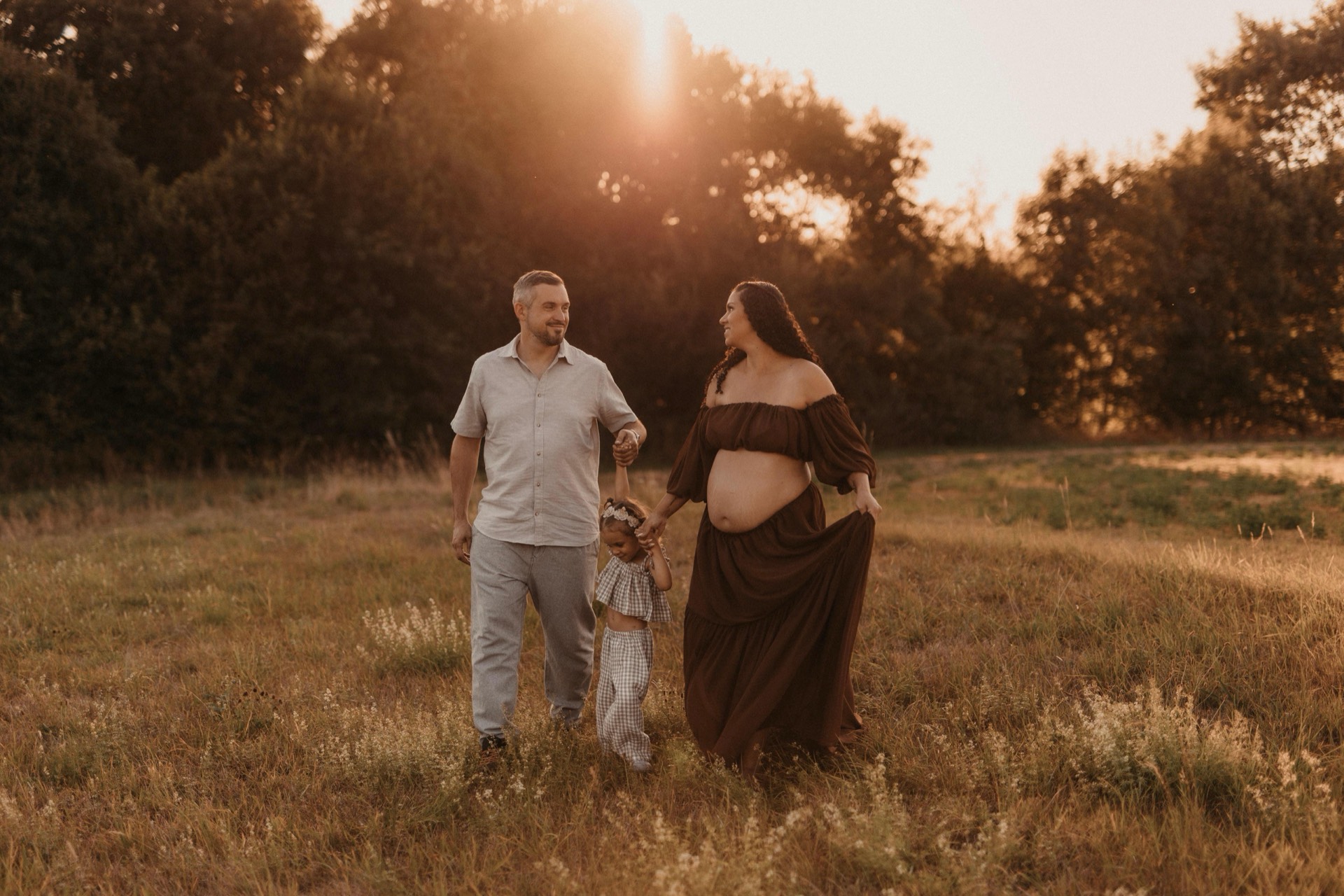 Familie spaziert Hand in Hand über eine sommerliche Wiese in der Abendsonne, die Mama erwartet ein Baby -- Familienfotografie von Simple Moments